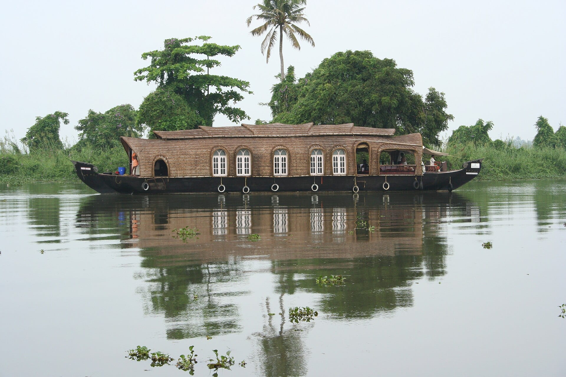 A houseboat moving through a narrow palm-lined backwater channel